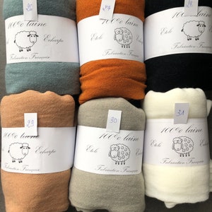 Pode incluir: Uma cole&ccedil;&atilde;o de cachec&oacute;is de l&atilde; enrolados em v&aacute;rias cores, incluindo verde-azulado, laranja, preto, bege, cinza e creme. Cada cachecol &eacute; rotulado com informa&ccedil;&otilde;es sobre o material e a origem. Os r&oacute;tulos tamb&eacute;m apresentam uma ovelha de desenho animado.