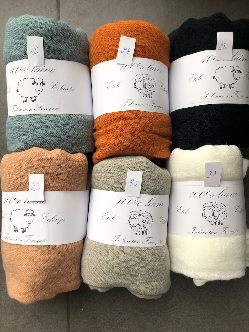 Pode incluir: Uma cole&ccedil;&atilde;o de cachec&oacute;is de l&atilde; enrolados em v&aacute;rias cores, incluindo verde-azulado, laranja, preto, bege, cinza e creme. Cada cachecol &eacute; rotulado com informa&ccedil;&otilde;es sobre o material e a origem. Os r&oacute;tulos tamb&eacute;m apresentam uma ovelha de desenho animado.