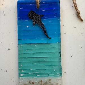 May include: A rectangular glass ornament with a whale shark design. The ornament features layers of blue and turquoise glass, with a white and tan beach-like base. A black and white whale shark is affixed to the front, and a jute string is attached for hanging.