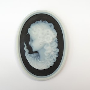 May include: Oval cameo pendant depicting a woman's profile with curly hair and a pearl necklace. The woman's image is light blue against a black background, framed by a light blue border. A classic piece of jewellery.