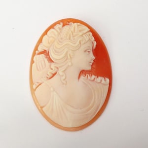 May include: Oval cameo pendant featuring a profile of a woman with flowing hair and a draped gown. The woman's image is carved in a cream color, set against an orange background. The cameo is a classic piece of jewelry.