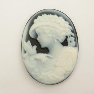 May include: Oval cameo pendant depicting a woman in profile, wearing a hat and holding a dove. The woman and dove are white against a black background, with a dark border. A classic, elegant design.