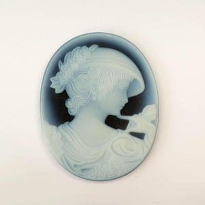 May include: Oval cameo pendant depicting a woman in profile, wearing a hat with floral detailing. The light blue figure is set against a dark blue backdrop, showcasing a classic design with intricate details.