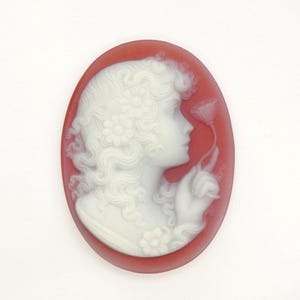 May include: Oval cameo pendant with a white profile of a woman with curly hair and flowers, holding a flower, set against a red background. A classic piece of jewellery.
