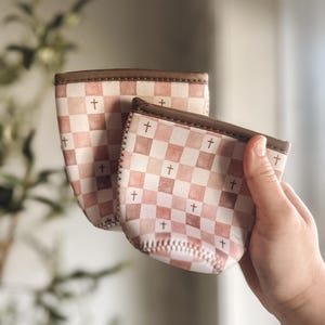 May include: Two neoprene beverage sleeves featuring a muted pink and white checkered pattern with small brown crosses printed on the squares. The sleeves have brown stitched trim along the top edges and a rounded base with visible white stitching.