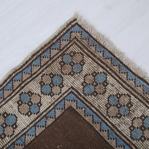 Puede incluir: Primer plano de una secci&oacute;n de una alfombra tejida con un patr&oacute;n geom&eacute;trico. La alfombra presenta un campo central marr&oacute;n, bordeado por una banda crema con dise&ntilde;os florales y en zigzag azules y marrones. Los bordes tienen un acabado con flecos.