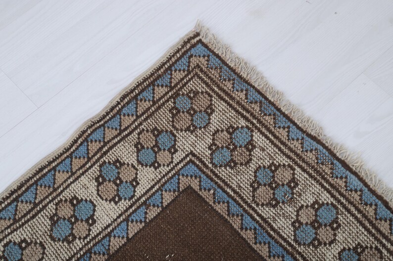Puede incluir: Primer plano de una secci&oacute;n de una alfombra tejida con un patr&oacute;n geom&eacute;trico. La alfombra presenta un campo central marr&oacute;n, bordeado por una banda crema con dise&ntilde;os florales y en zigzag azules y marrones. Los bordes tienen un acabado con flecos.
