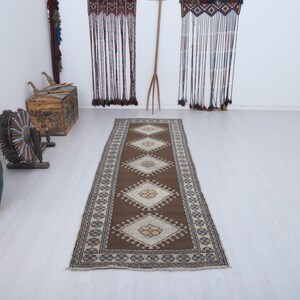 Puede incluir: Una alfombra rectangular marr&oacute;n con un patr&oacute;n central de diamantes y un borde de formas geom&eacute;tricas repetidas. La alfombra est&aacute; colocada sobre un suelo de color claro, con otros objetos decorativos en el fondo.