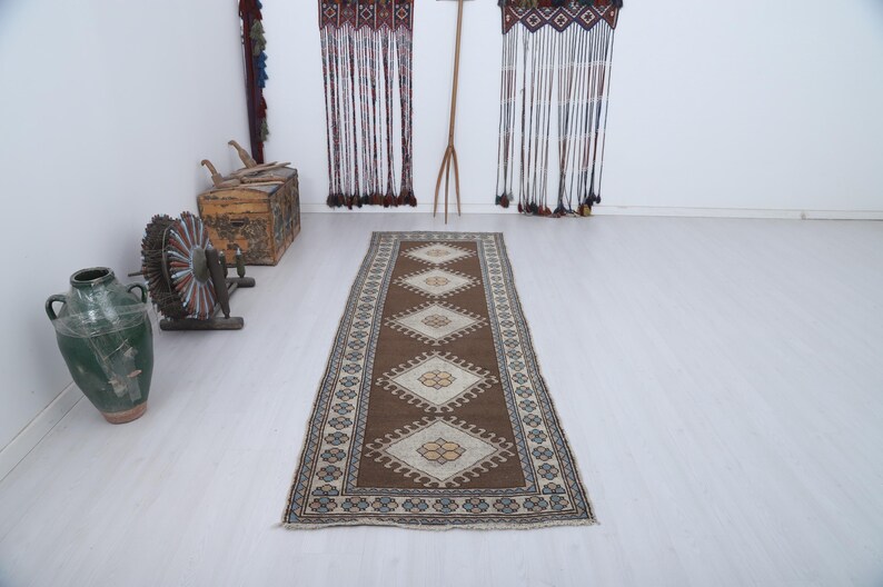 Puede incluir: Una alfombra rectangular marr&oacute;n con un patr&oacute;n central de diamantes y un borde de formas geom&eacute;tricas repetidas. La alfombra est&aacute; colocada sobre un suelo de color claro, con otros objetos decorativos en el fondo.