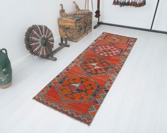 May include: An orange runner rug with geometric patterns in shades of orange, brown, and gray. The rug has a rectangular shape and is placed on a white floor. The rug's design features repeating diamond and star-like motifs.
