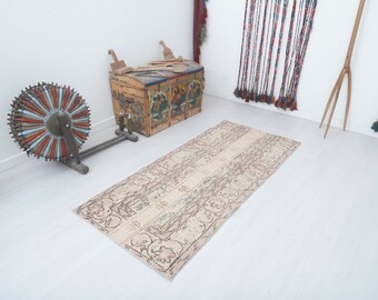 Puede incluir: Una alfombra vintage de color crema con un patrón floral y un aspecto descolorido y desgastado. La alfombra está colocada sobre un suelo de madera blanco.
