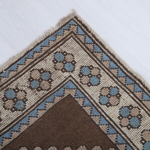 Puede incluir: Primer plano de una esquina de una alfombra estampada marr&oacute;n, beige y azul. La alfombra presenta un dise&ntilde;o geom&eacute;trico con patrones florales y triangulares repetidos. La alfombra tiene un borde con flecos.