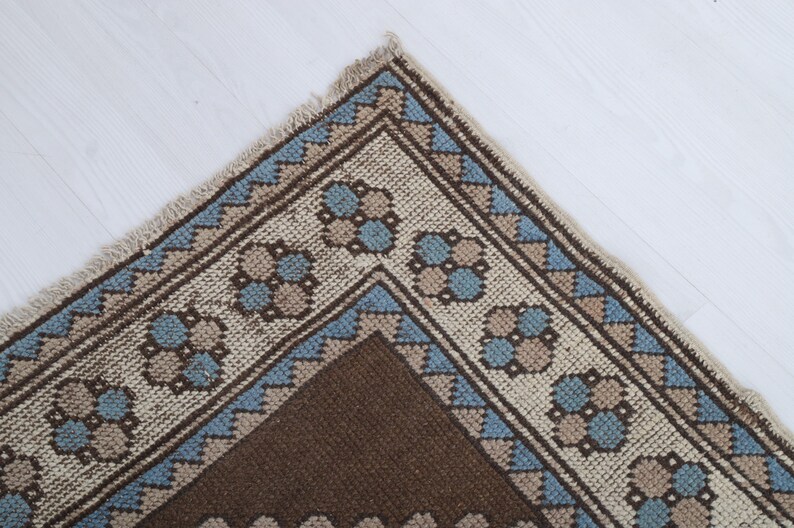 Puede incluir: Primer plano de una esquina de una alfombra estampada marr&oacute;n, beige y azul. La alfombra presenta un dise&ntilde;o geom&eacute;trico con patrones florales y triangulares repetidos. La alfombra tiene un borde con flecos.