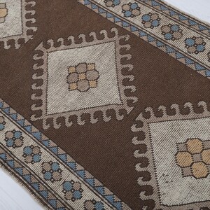 Puede incluir: Una alfombra estampada marr&oacute;n y beige con un dise&ntilde;o central de diamante. La alfombra presenta patrones geom&eacute;tricos repetidos en marr&oacute;n, beige y azul. El borde de la alfombra tiene un patr&oacute;n floral repetido.