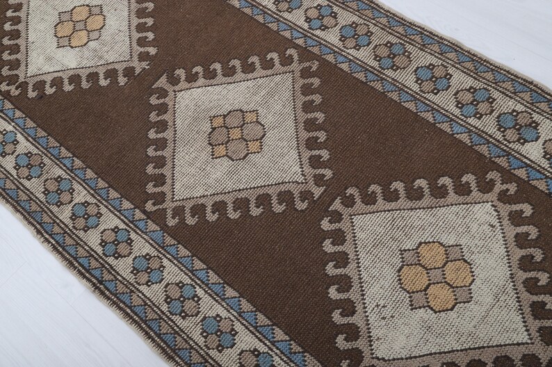 Puede incluir: Una alfombra estampada marr&oacute;n y beige con un dise&ntilde;o central de diamante. La alfombra presenta patrones geom&eacute;tricos repetidos en marr&oacute;n, beige y azul. El borde de la alfombra tiene un patr&oacute;n floral repetido.