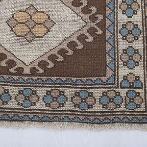 Puede incluir: Primer plano de una secci&oacute;n de una alfombra estampada marr&oacute;n, beige y azul. La alfombra presenta un dise&ntilde;o central de diamante con un motivo floral, rodeado por un borde con patrones florales y geom&eacute;tricos repetidos. La alfombra tiene un borde con flecos.