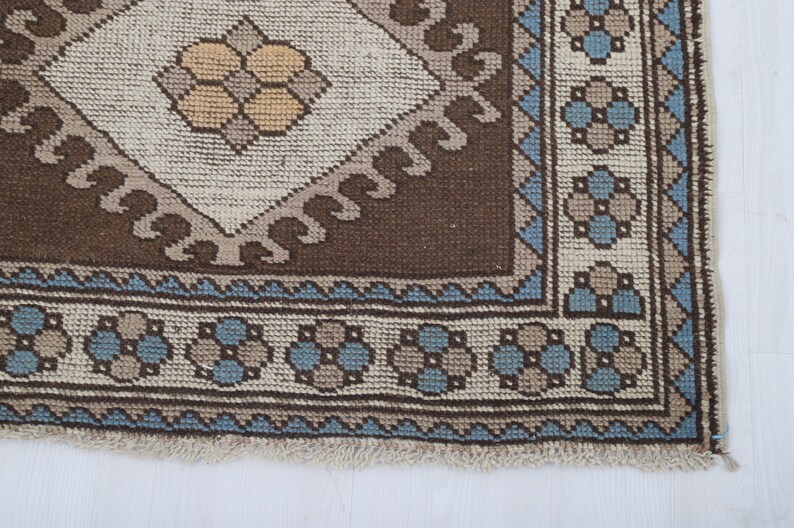 Puede incluir: Primer plano de una secci&oacute;n de una alfombra estampada marr&oacute;n, beige y azul. La alfombra presenta un dise&ntilde;o central de diamante con un motivo floral, rodeado por un borde con patrones florales y geom&eacute;tricos repetidos. La alfombra tiene un borde con flecos.