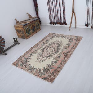 May include: Rectangular rug with a cream-colored center and a pink floral border. The rug features a central medallion design and is surrounded by a detailed floral pattern in shades of pink, green, and brown. The rug is placed on a light-colored wooden floor.