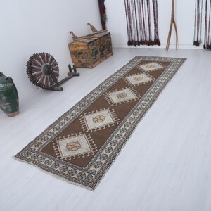 Puede incluir: Una alfombra larga y rectangular de color marr&oacute;n con un patr&oacute;n de diamantes repetido en crema y beige. La alfombra tiene un borde con un dise&ntilde;o geom&eacute;trico azul y blanco. La alfombra probablemente se usa como corredor de pasillo.