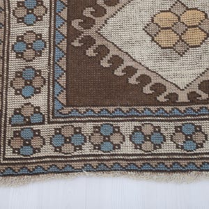 Puede incluir: Primer plano de una secci&oacute;n de una alfombra vintage con un patr&oacute;n geom&eacute;trico. La alfombra presenta un dise&ntilde;o central de diamante en beige y marr&oacute;n, rodeado por un borde marr&oacute;n y un patr&oacute;n floral azul y beige. La alfombra tiene un borde con flecos.