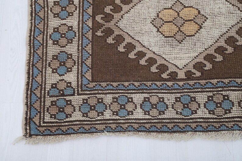 Puede incluir: Primer plano de una secci&oacute;n de una alfombra vintage con un patr&oacute;n geom&eacute;trico. La alfombra presenta un dise&ntilde;o central de diamante en beige y marr&oacute;n, rodeado por un borde marr&oacute;n y un patr&oacute;n floral azul y beige. La alfombra tiene un borde con flecos.