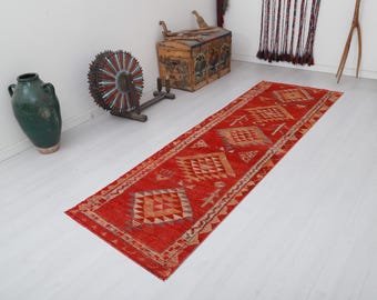 May include: A red, orange, and cream colored runner rug with a geometric pattern. The rug has a traditional design with repeating diamond shapes and triangles.