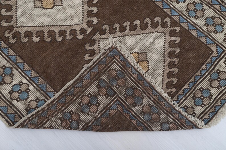 Puede incluir: Primer plano de una alfombra estampada marr&oacute;n y beige con un dise&ntilde;o geom&eacute;trico. La alfombra presenta un patr&oacute;n repetitivo de cuadrados y motivos florales en tonos marrones, beiges y azules. El borde de la alfombra tiene un borde decorativo.