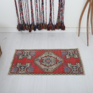 May include: A small, rectangular red rug with a central geometric design in beige, blue, and gray. The rug features a border and repeating patterns in shades of blue and gray. The rug is approximately 24 inches long and 12 inches wide.
