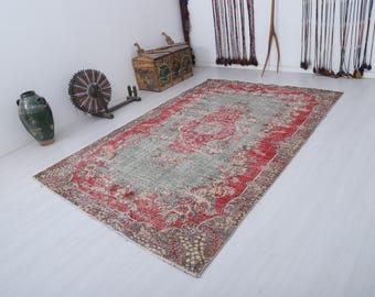 May include: A rectangular, distressed-look rug with a central floral design in red and a faded green background. The rug has a red border and a beige, patterned edge. Other items in the image include a green vase, a wooden chest, and a decorative wheel.
