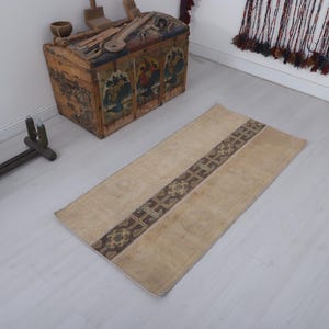 May include: A rectangular area rug featuring a light beige field and a central vertical band decorated with a dark brown geometric pattern. The rug has a low pile texture and is positioned flat on a light-colored floor surface.
