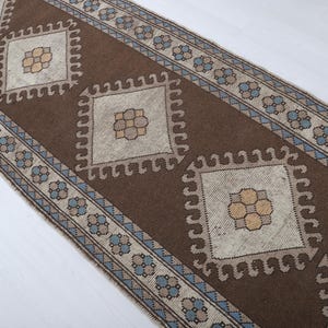 Puede incluir: Una alfombra de pasillo marr&oacute;n y beige con un patr&oacute;n geom&eacute;trico. La alfombra presenta formas de diamante repetidas con dise&ntilde;os florales centrales. El borde tiene un patr&oacute;n floral y geom&eacute;trico repetido en azul y beige.