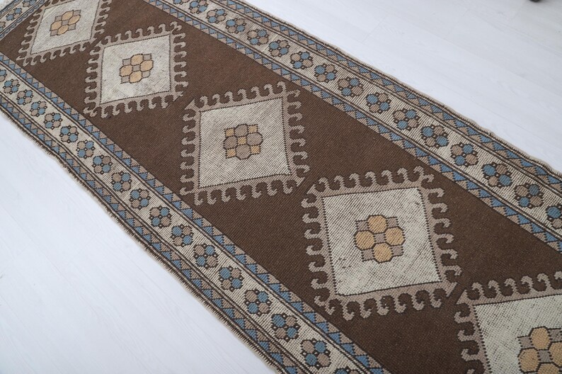 Puede incluir: Una alfombra de pasillo marr&oacute;n y beige con un patr&oacute;n geom&eacute;trico. La alfombra presenta formas de diamante repetidas con dise&ntilde;os florales centrales. El borde tiene un patr&oacute;n floral y geom&eacute;trico repetido en azul y beige.