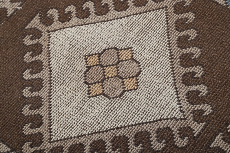 Puede incluir: Primer plano de una alfombra tejida con un dise&ntilde;o cuadrado central. La alfombra presenta un patr&oacute;n repetitivo de colores marr&oacute;n, beige y tostado. El cuadrado central tiene un dise&ntilde;o floral en el centro. La alfombra tiene una superficie texturizada.