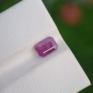 Puede incluir: Una gema de color rojo púrpura, tallada en forma rectangular. La gema se encuentra sobre una superficie blanca texturizada, con un fondo borroso verde y rojo. Las facetas de la gema reflejan la luz.