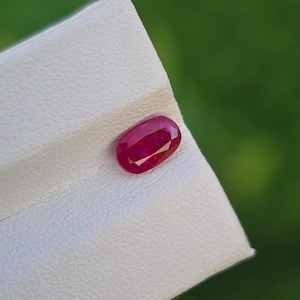 May include: A faceted, oval-shaped ruby gemstone, displaying a deep red hue. The gem is set against a textured white surface, with a blurred green background. The ruby's polished facets reflect light, showcasing its clarity and brilliance.