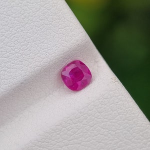 May include: A cushion-cut, vibrant pink ruby gemstone. The gemstone is set against a white textured surface, with a blurred green background. The ruby's facets reflect light, showcasing its brilliance.