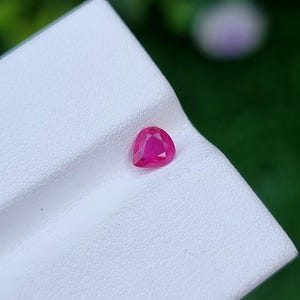 May include: A triangular-cut ruby gemstone in a vivid pink-red hue. The gem is displayed on a white, textured surface. The background is a soft, blurred green.