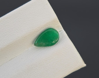 May include: A teardrop-shaped, polished green gemstone rests on a white surface against a dark gray background. The gemstone has a smooth, reflective surface and a vibrant, emerald green color. The stone is likely a cabochon, suitable for jewelry.