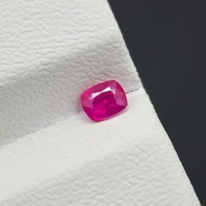 May include: A cushion-cut ruby gemstone, displaying a rich, red hue. The gem's facets reflect light, creating a sparkling effect. The ruby is presented on a textured white surface, with a dark backdrop.