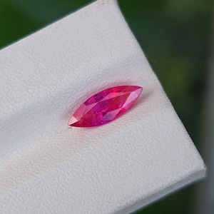 Könnte beinhalten: Ein facettierter Rubin im Marquiseschliff, der einen leuchtenden rosa-roten Farbton aufweist. Der Edelstein liegt auf einer weißen Oberfläche, mit einem verschwommenen grünen Hintergrund. Die längliche Form und die Facetten erzeugen einen funkelnden Effekt.