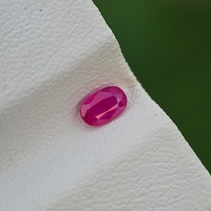 May include: A faceted, oval-shaped ruby gemstone. The ruby is a vibrant shade of red and is set against a textured white surface. The background is a soft green, providing a contrast to the gemstone's color. This image showcases a precious stone.
