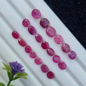 May include: A collection of oval-cut, vibrant red rubies arranged on a white surface. The gemstones vary slightly in size and shade, showcasing a range of rich, crimson hues. A sprig of purple flowers adds a touch of color.