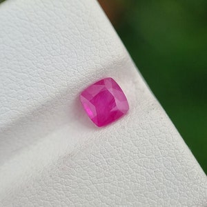 May include: A vibrant, cushion-cut ruby gemstone. The gem is a deep pinkish-red color, with facets that catch the light. The ruby is set against a white textured surface, with a blurred green background.