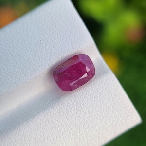 Könnte beinhalten: Ein facettierter, oval geformter Rubin-Edelstein. Der Rubin hat eine tiefe, leuchtende rote Farbe mit sichtbaren inneren Einschlüssen. Er liegt auf einer weißen, strukturierten Oberfläche mit einem verschwommenen grünen Hintergrund.