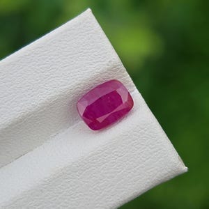 Puede incluir: Una gema de rubí con corte cojín, que muestra un tono rojo intenso. La gema se presenta sobre una superficie blanca texturizada, con un fondo verde borroso. Las facetas del rubí capturan la luz, mostrando su brillo.