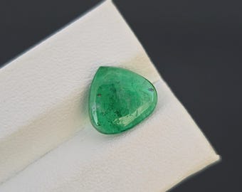 May include: A triangular, polished emerald gemstone with a vibrant green hue. The gem is set against a white surface, with a dark gray background. The emerald's surface reflects light, showcasing its clarity and color.