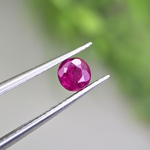 May include: A round, faceted, deep pinkish-red gemstone held by a pair of silver-toned metal tweezers. The stone features a brilliant cut with multiple triangular facets that reflect light, showcasing a vibrant, saturated color throughout the gem.
