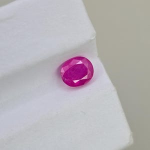 以下が含まれることがあります： ファセット加工された、楕円形のルビーの宝石。この宝石は鮮やかで、深いピンクがかった赤色をしており、内部のファセットが見えます。ルビーは白い背景に置かれています。