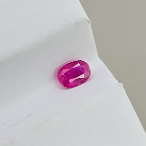 May include: A faceted, oval-shaped ruby gemstone. The gem is a vibrant shade of pinkish-red, with visible internal facets that reflect light. The ruby is set against a white background.