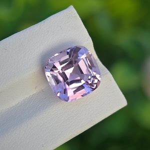 May include: A faceted, light purple gemstone with a square cushion cut. The gem is held on a white surface, with a blurred green background. The gem's facets reflect light, creating a sparkling effect.